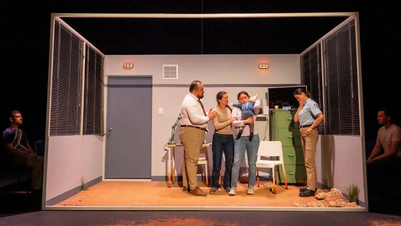 Un grupo de actores de teatro apárecen en una escenografía que representa una oficina de migración.